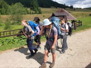 Štyri dievčatá v skautských šatkách kráčajú s veľkými turistickými batohmi po štrkovej ceste v horskej prírode