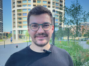 Usmievajúci sa muž v okuliaroch a čiernom tričku s klipovým mikrofónom stojí v mestskom parku pred výškovou budovou.