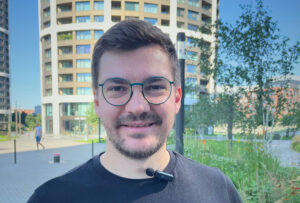 Usmievajúci sa muž v okuliaroch a čiernom tričku s klipovým mikrofónom stojí v mestskom parku pred výškovou budovou.